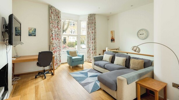 A modern living room with a wooden floor, a grey sofa, a blue armchair, a desk with a computer, and a large window with floral curtains. - Elegant 4-Bed Islington Home with Garden (London)