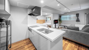 Uma cozinha moderna com uma ilha central, máquina de lavar louça embutida e um sofá cinzento na sala de estar.