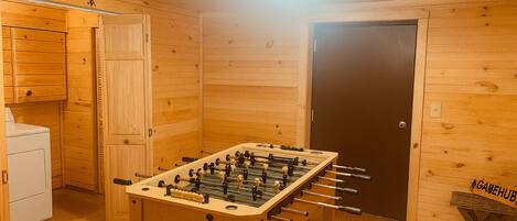 Habitació amb interior de fusta, taula de futbolí, zona de bugaderia i una porta amb la inscripció 'GAMEHUB'.