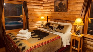 A log cabin room with a bed, two bedside lamps, a framed picture on the wall, and a window with curtains. - Crabapple: Charming cabin with hot tub, pond, creek access in Blue Ridge (Morganton)