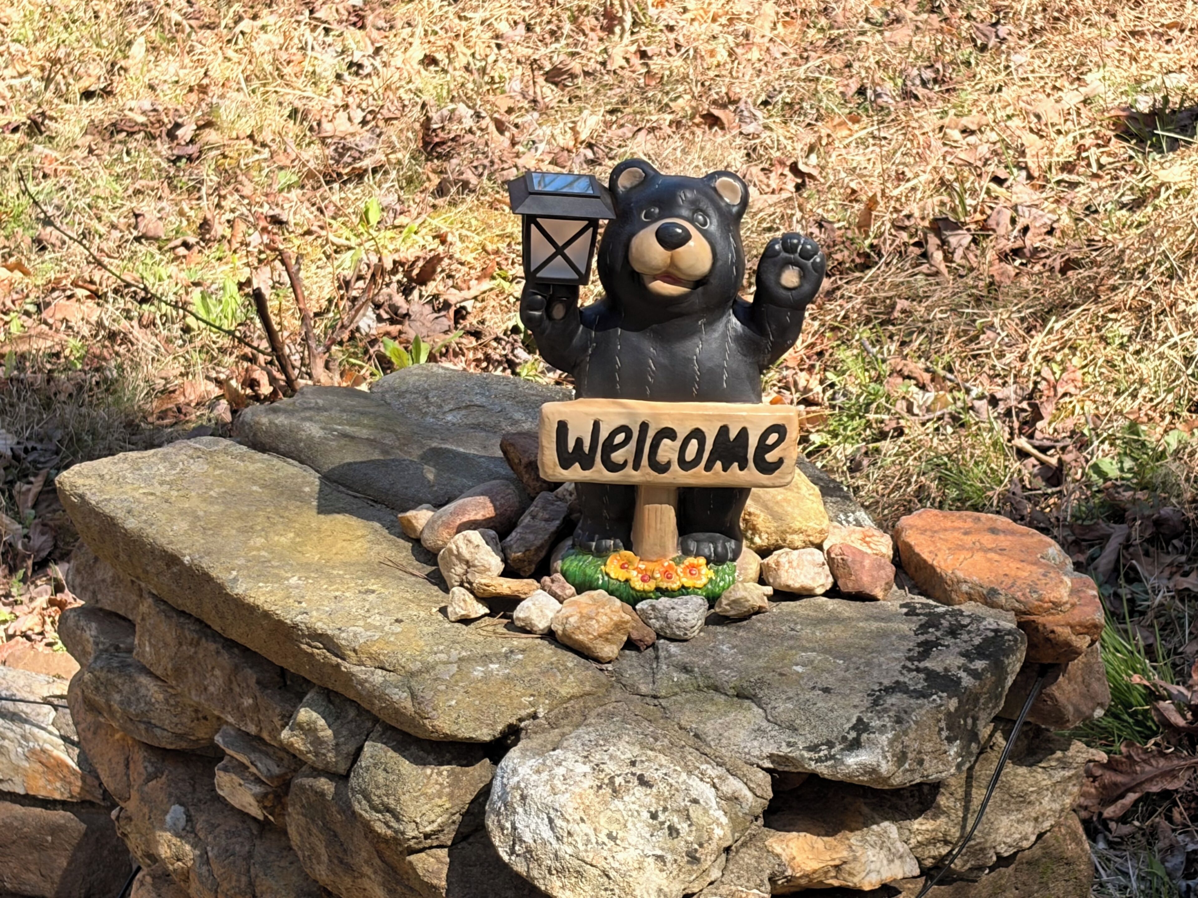 A bear statue holding a welcome sign, surrounded by rocks and foliage.