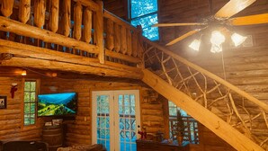 A rustic cabin interior with a wooden staircase, a ceiling fan, a television, and a large window with a scenic view. - Crabapple: Charming cabin with hot tub, pond, creek access in Blue Ridge (Morganton)