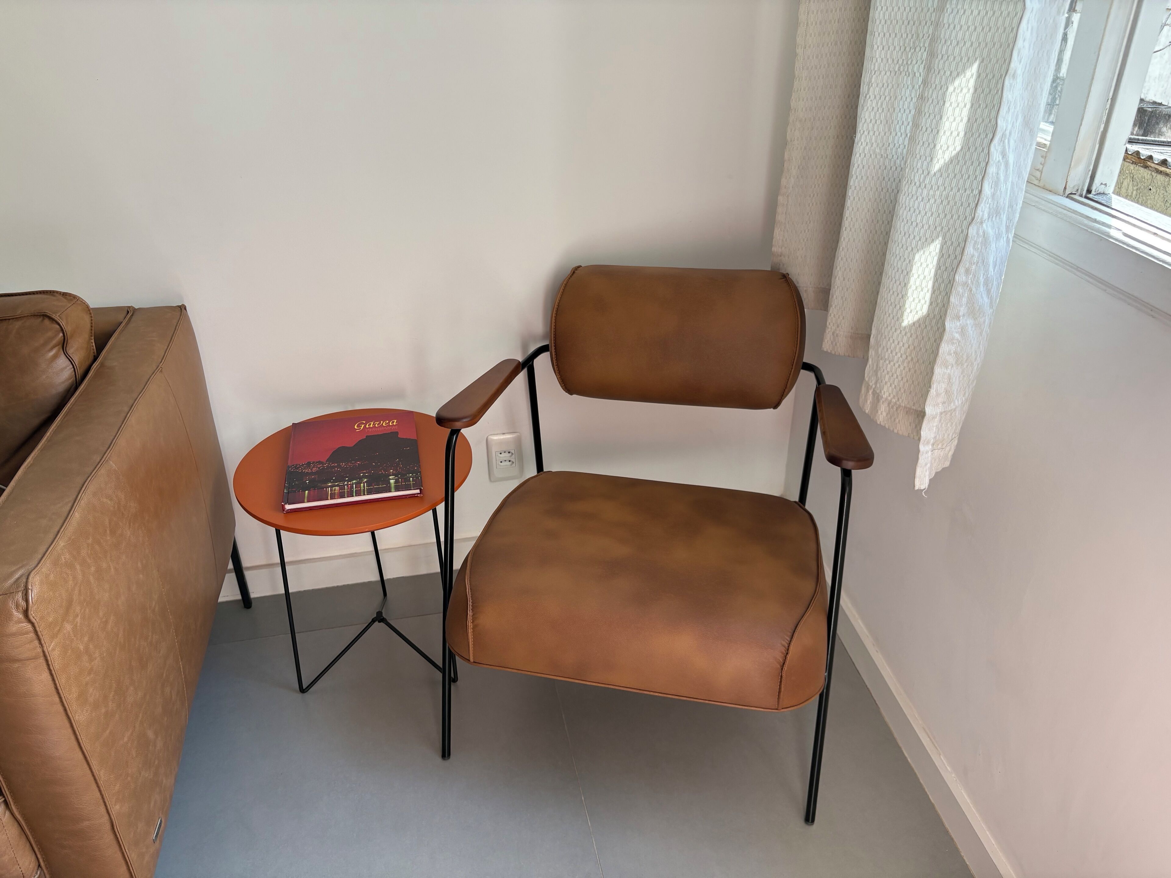 A brown leather chair with a small round table beside it, a book titled 'Gavea' on the table, and a window with a white curtain.