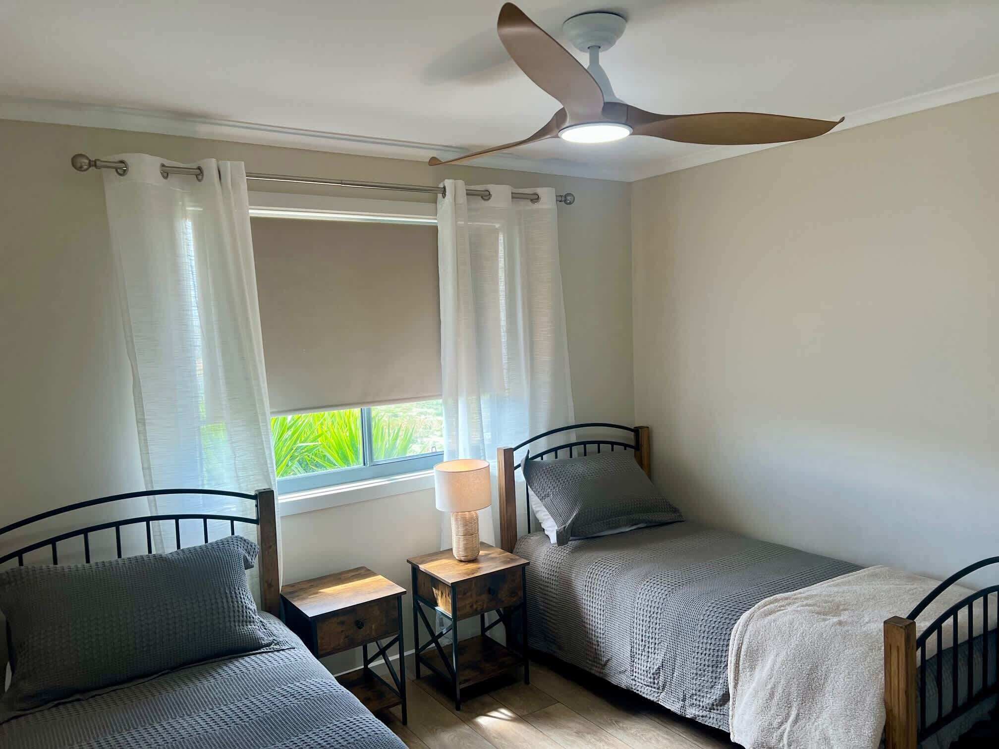 A bedroom with two single beds, a ceiling fan, a bedside table with a lamp, and a window with curtains.