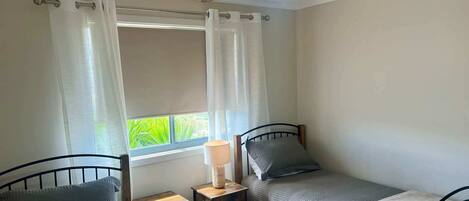 A bedroom with two single beds, a ceiling fan, a bedside table with a lamp, and a window with curtains.