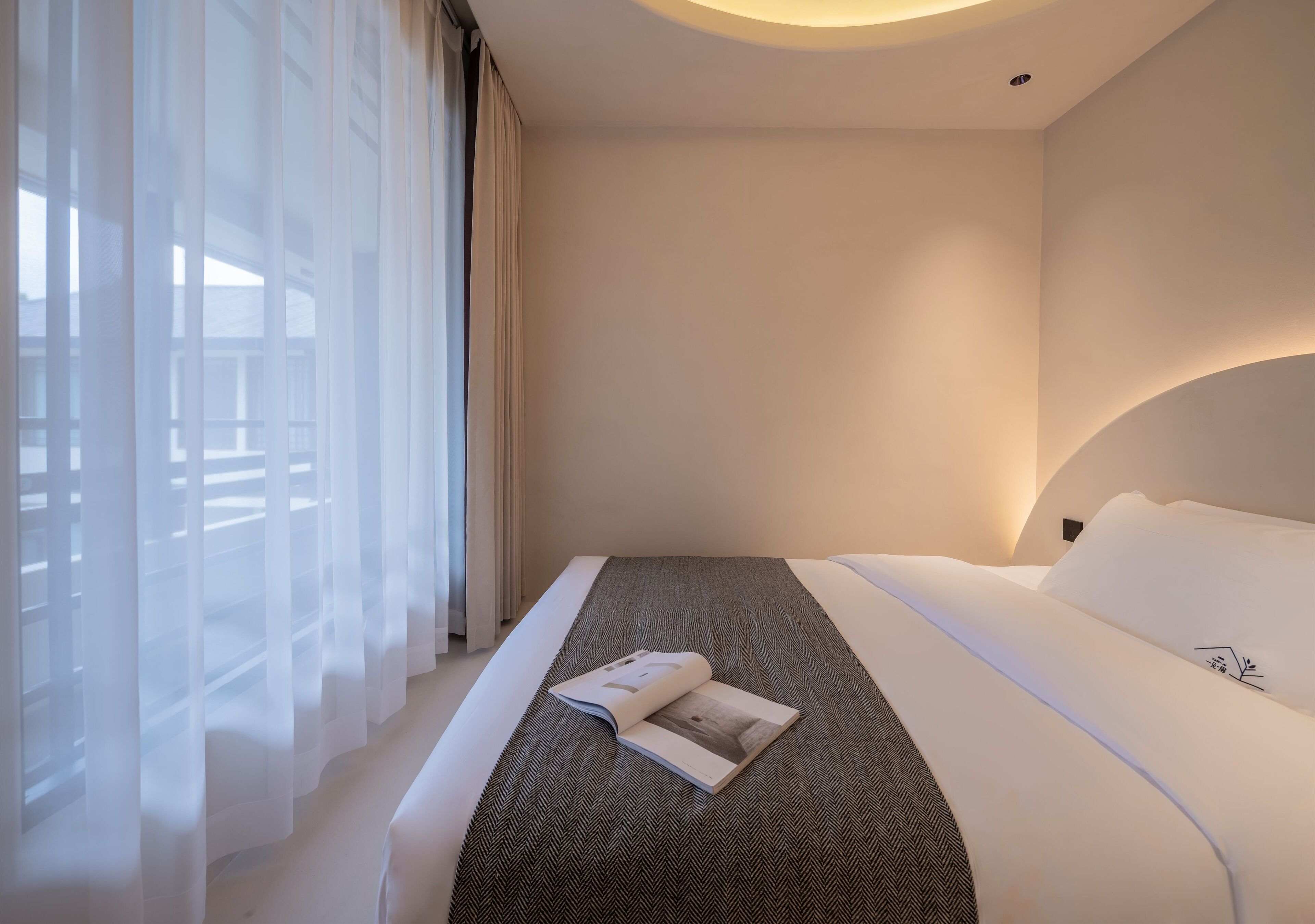 A hotel room with a bed, a magazine on the bed, and sheer curtains on the window.