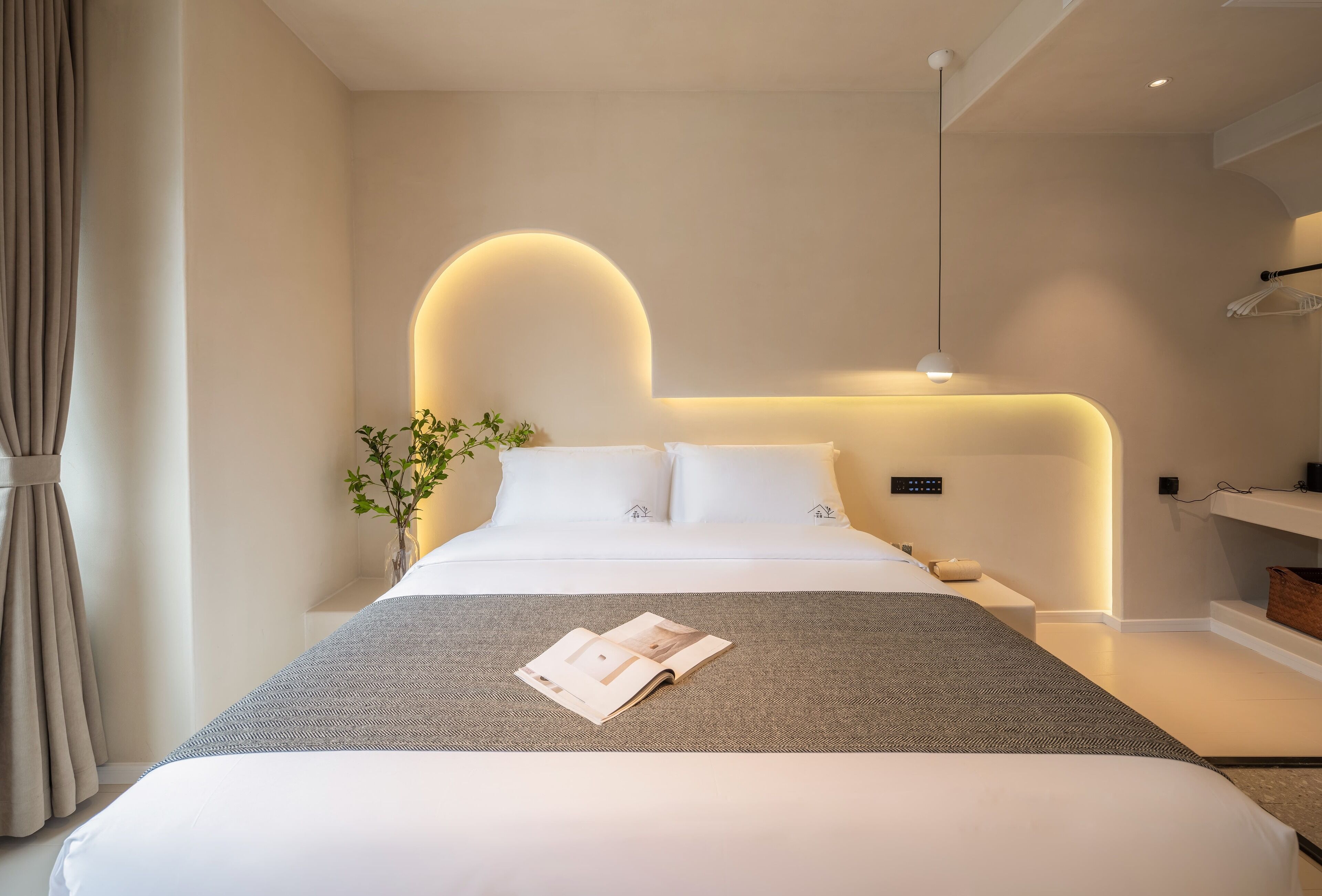 A modern hotel room with a large bed, a bedside table, a wall-mounted light, and a plant.