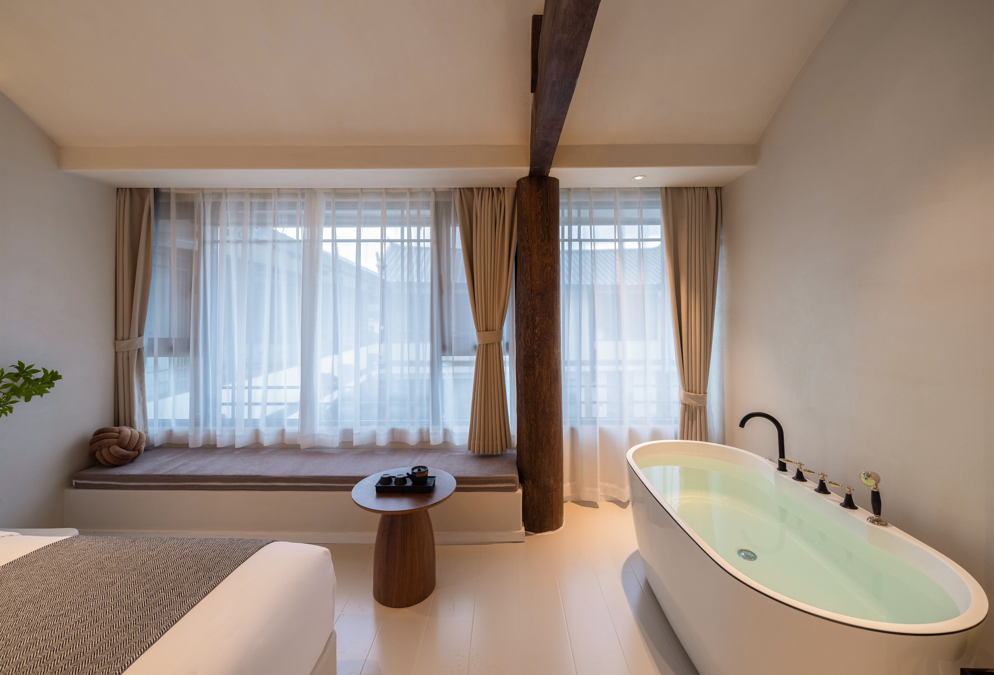 Een moderne badkamer met een vrijstaand bad, een raamzitje met gordijnen en een kleine ronde tafel met een decoratief object.
