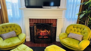 Een woonkamer met een open haard, twee groene fauteuils en een televisie boven de haardmantel.
