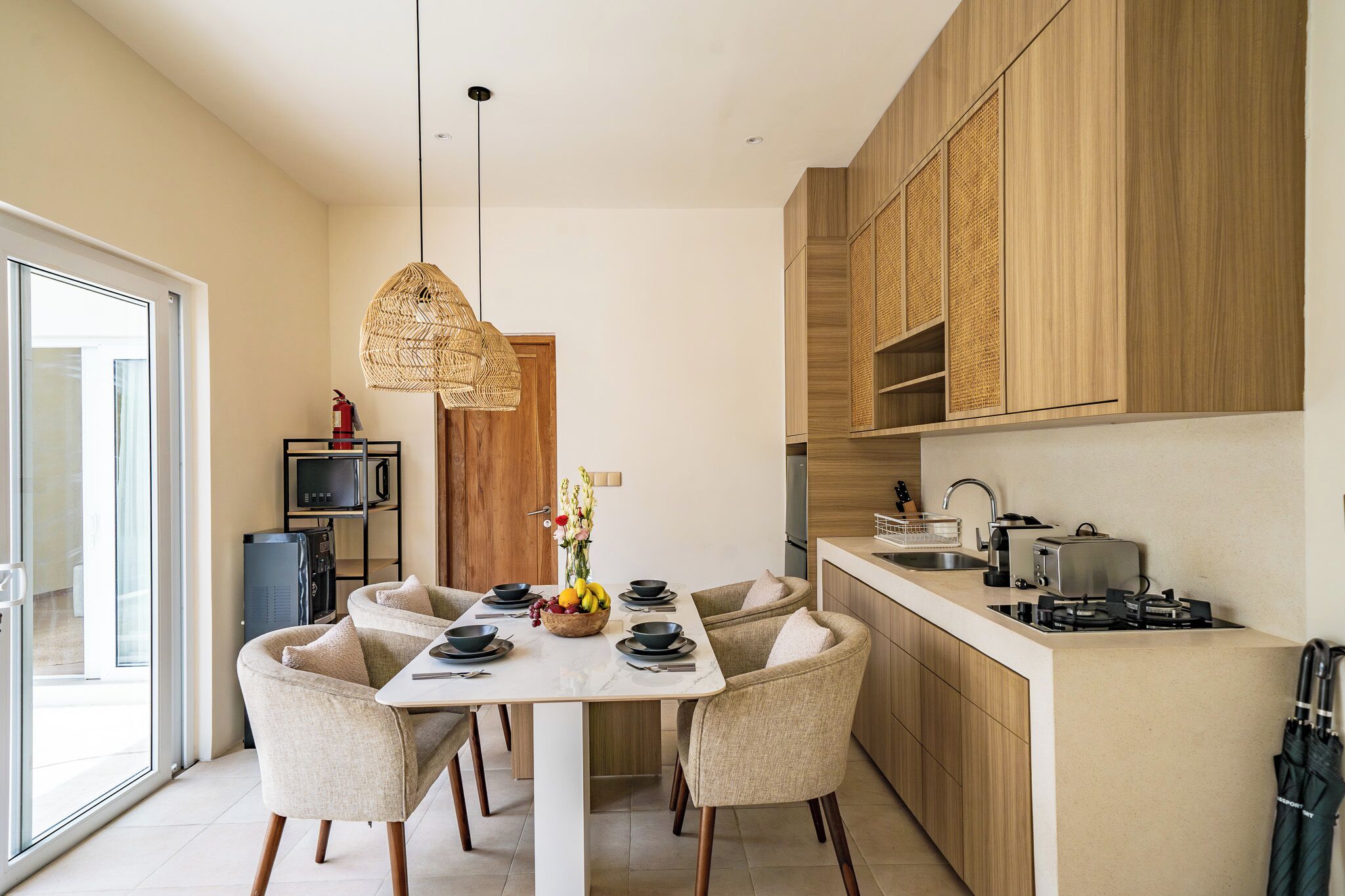 A modern kitchen with a dining area, featuring a white table, beige chairs, and a woven pendant light.