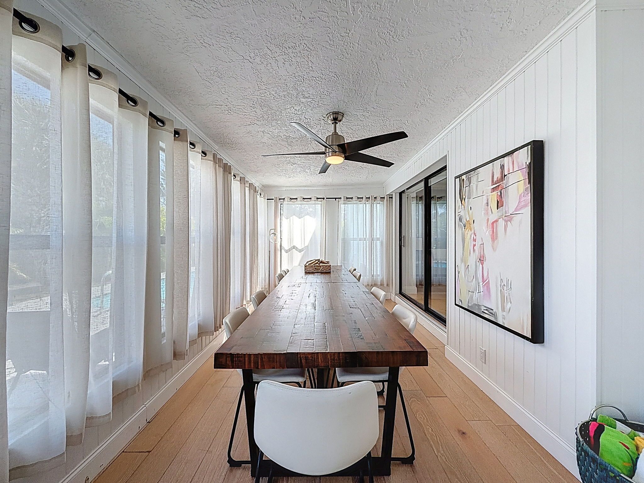 Une longue table à manger entourée de chaises, un ventilateur de plafond et de grandes fenêtres avec des rideaux.
