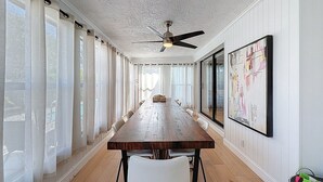 A long dining table with chairs, a ceiling fan, and large windows with curtains.
