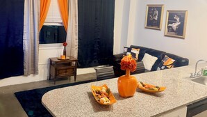 Ein modernes Wohnzimmer mit Küchenbereich, ausgestattet mit einem Esstisch mit Obst und einer Vase, einer schwarzen Ledersofa und einem Fenster mit Vorhängen.