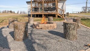 A wooden deck with a fire pit surrounded by tree stumps, a covered outdoor seating area, and a gravel ground. - Charming 2 Bedroom 2 Bath Cottage near the lake. (Campbellsville)