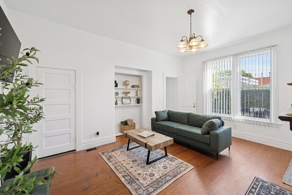 Ein Wohnzimmer mit einem grünen Sofa, einem Holztisch und einem gemusterten Teppich. Eine weiße Tür, ein Regal mit Dekoration und ein Fenster mit Jalousien sind ebenfalls zu sehen.