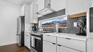 Una cocina moderna con armarios blancos, electrodomésticos de acero inoxidable, un amplio ventanal con vistas a la montaña y un salpicadero con diseño geométrico.