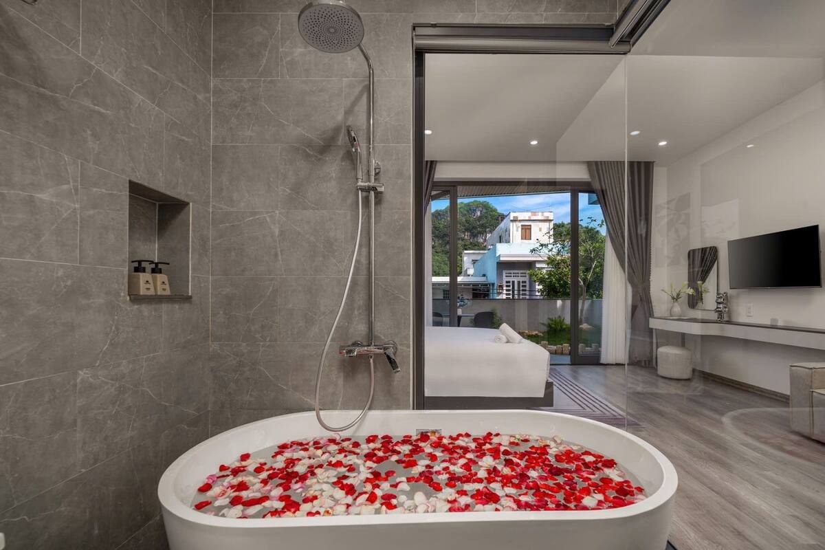A modern bathroom with a freestanding bathtub filled with rose petals, a showerhead, and a large window offering a view of the outside.