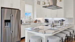 A modern kitchen with a stainless steel refrigerator, white cabinetry, a large island with bar stools, and a window providing a view. - Live in Luxury at This Brand New Tybee Island Escape | Heated Pool Access (Tybee Island)