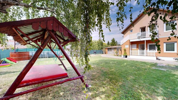 A backyard with a swing set, a grassy lawn, a wooden fence, and a two-story house with a balcony. - Holiday apartment with pool, whirlpool, sauna and hot tub (Balatonszabadi)