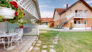 A courtyard with a white outdoor seating area, a white lattice structure, and a two-story building with a red roof and white fence. - Holiday apartment with pool, whirlpool, sauna and hot tub (Balatonszabadi)