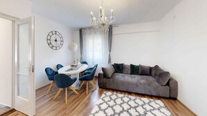 A living room with a grey sofa, blue chairs, a round table, a chandelier, and a large wall clock. - Holiday apartment with pool, whirlpool, sauna and hot tub (Balatonszabadi)