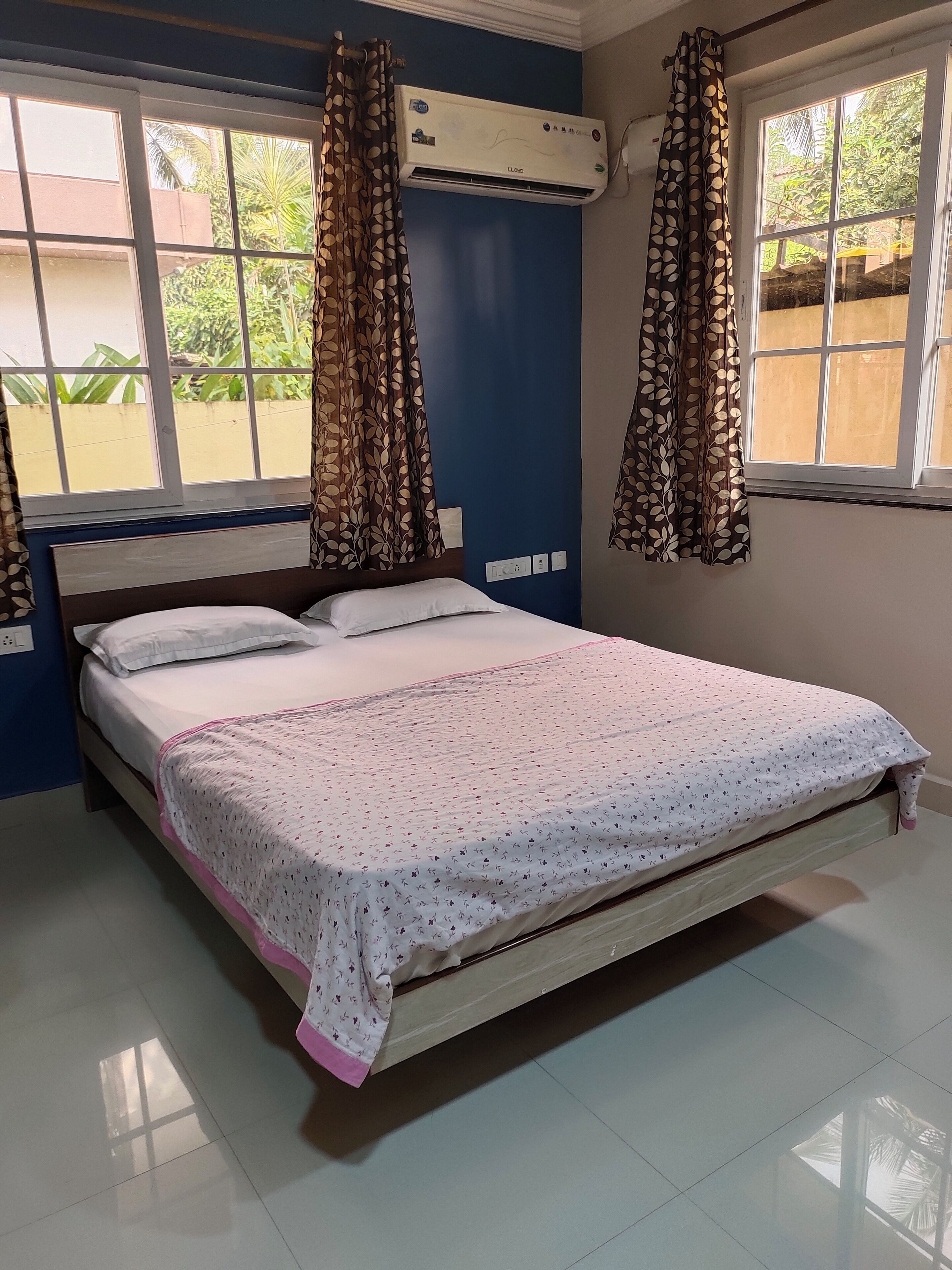 A bedroom with a bed, curtains, an air conditioning unit, and a window with a view of greenery.
