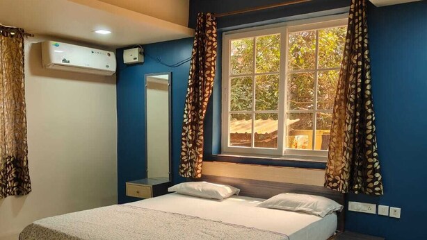 A bedroom with a bed, a ceiling fan, an air conditioning unit, a window with curtains, and a nightstand. - Peaceful 2-bedroom apartment in charming Colva perfect for relaxing (Fatona)