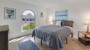 Un dormitorio con una cama grande, un escritorio, una silla, un ventanal con vista a un estacionamiento y un cuadro en la pared.