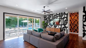 A modern living room with a grey sofa, a ceiling fan, and a large window overlooking a patio area. - Contour - Priv Pool- Fun Design- Mins 2 everything (Phoenix)
