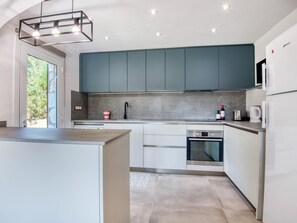 A modern kitchen with a central island, built-in appliances, and a large window. - Villa Florència: Your new exclusive option in Calonge - Costa Brava. (Calonge i Sant Antoni)