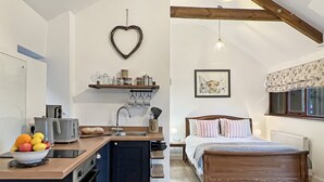 A compact, modern room with a kitchenette, a bed with a wooden headboard, a hanging light fixture, and a window with a patterned valance. - Daveys Barn, Brendon Hill on Exmoor (Nr Watchet)