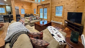 L'intérieur d'un chalet confortable, avec une salle à manger, un salon doté de canapés et d'un téléviseur, et de grandes fenêtres offrant une vue sur l'extérieur.