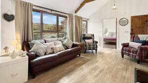 A cozy living room with a brown leather sofa, plaid armchairs, and a wooden floor. A window with curtains offers a view of the outdoors. - Westlands, Brendon Hill on Exmoor (Nr Watchet)