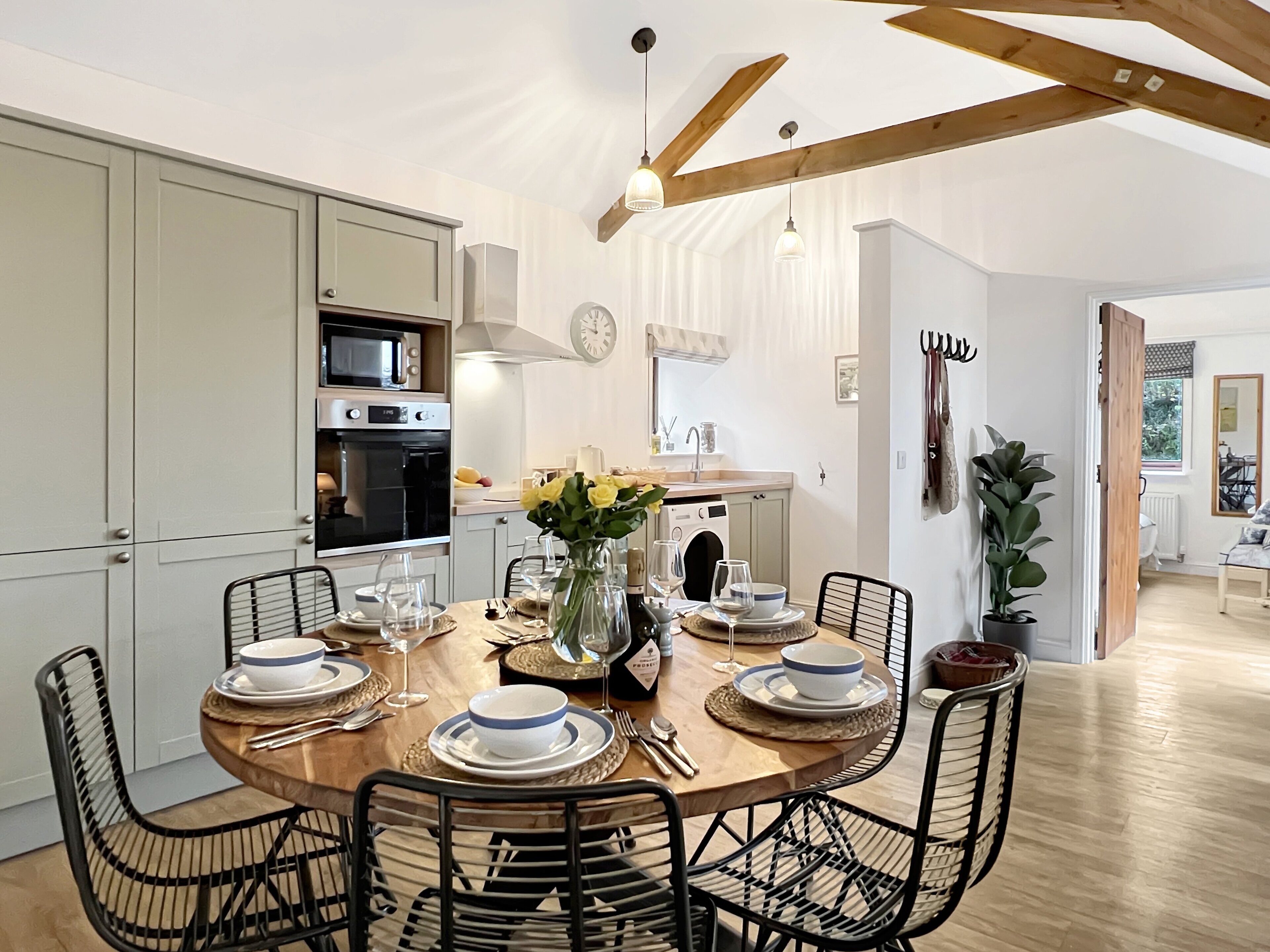 A modern kitchen with a dining area, featuring wooden beams, a round table set for six, and a view into the living room.