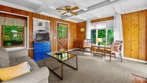A living room with wooden walls, a ceiling fan, a blue dresser, a television, a coffee table, a sofa, and two chairs. - Tranquil Eastham Waterfront Cottage Escape (Eastham)