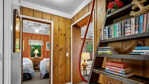 A room with wooden walls, a bookshelf, a bed with a patterned bedspread, and a window with a view of greenery. - Tranquil Eastham Waterfront Cottage Escape (Eastham)