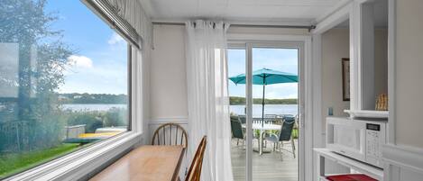 A room with a wooden table and chairs, a sliding glass door leading to an outdoor area with a table and chairs, and a view of a body of water.