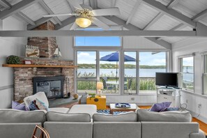 A spacious living room with a fireplace, a television, and a large window overlooking a lake. - Charming Waterfront Escape Along the Cove (Eastham)