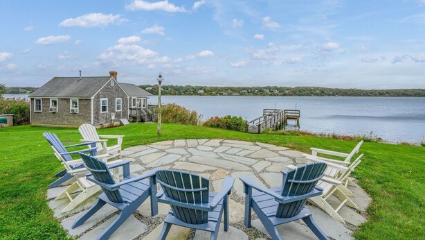 A stone patio with Adirondack chairs, a house with a dock, and a lake view.
