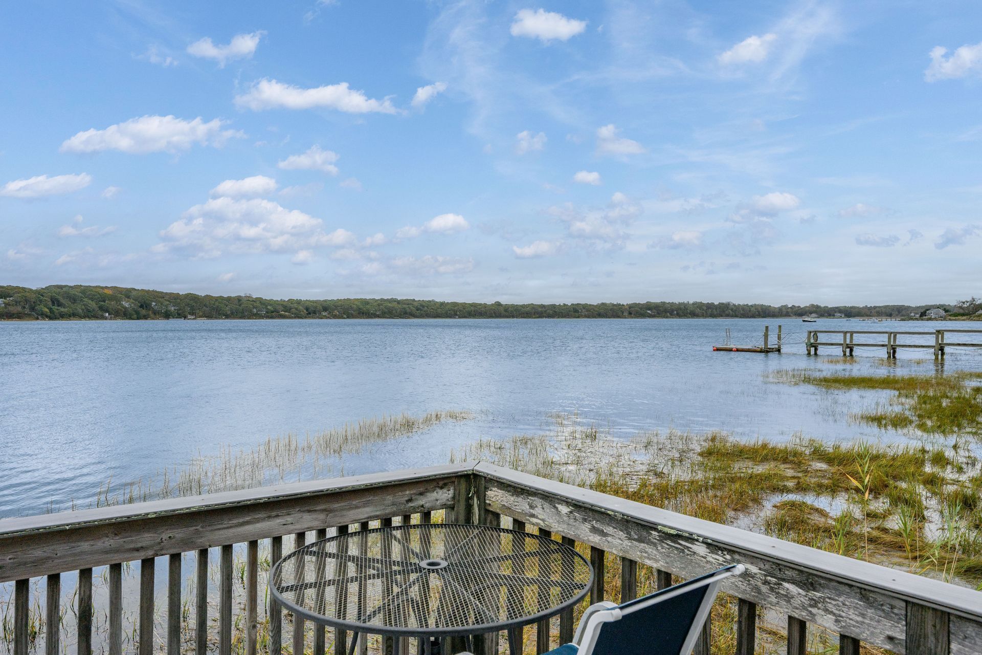 A wooden deck with a metal table and chairs overlooking a calm lake with a distant shoreline.