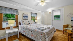 Un dormitori amb un llit, taules de nit, ventilador al sostre, finestra amb cortines, una porta i una petita cadira.
