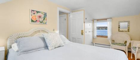 Un dormitorio con una cama grande, un cuadro de flores, una puerta que da a otra habitación y vista al mar por una ventana.