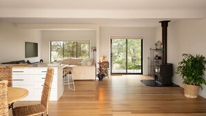 Amplia sala de estar con piso de madera, un sofá, una mesa de comedor con sillas, un televisor montado en la pared y un estufa de leña negra.