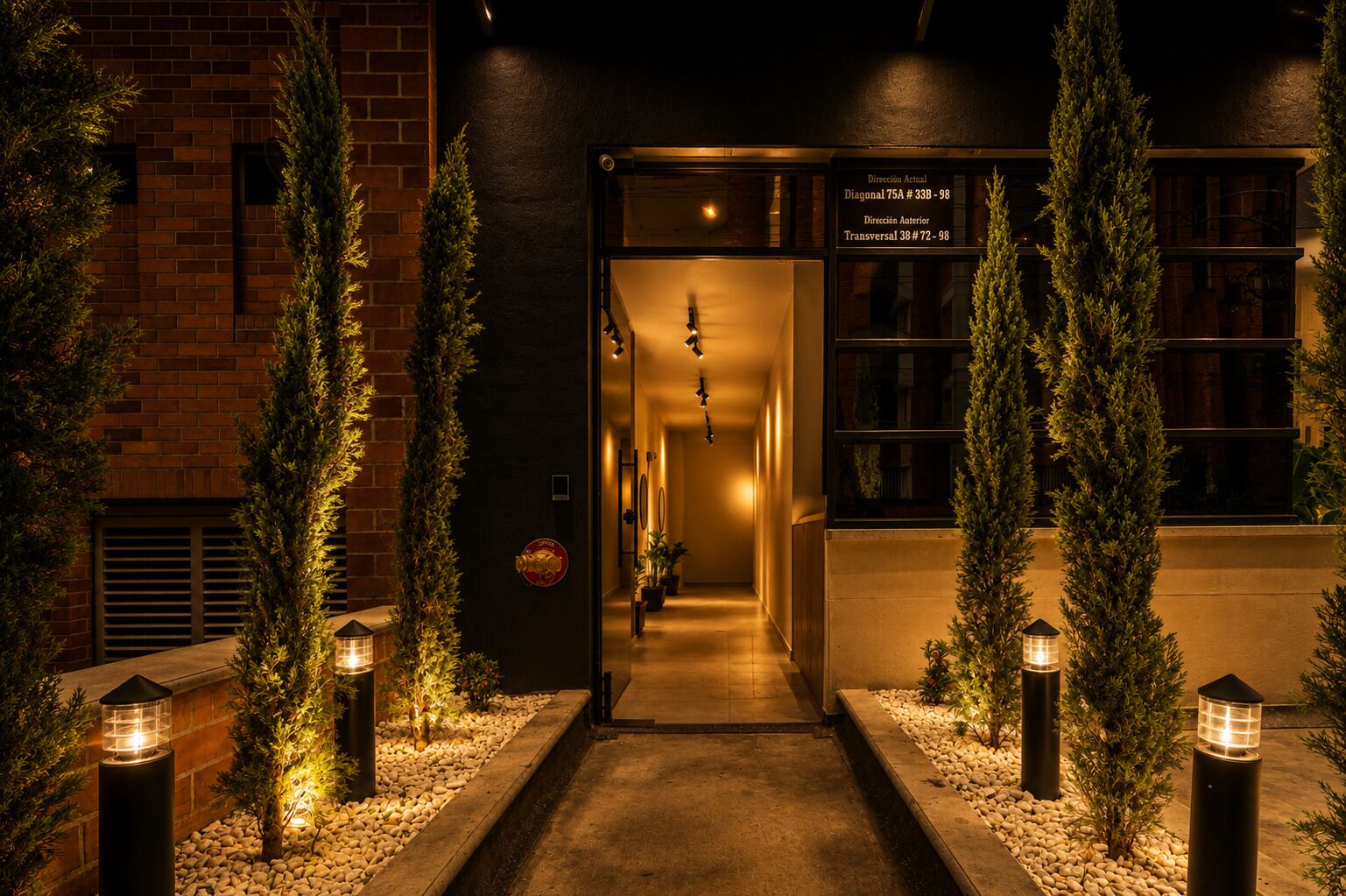 A hotel entrance with a brick facade, a sign indicating directions, and a pathway flanked by trees and lanterns.