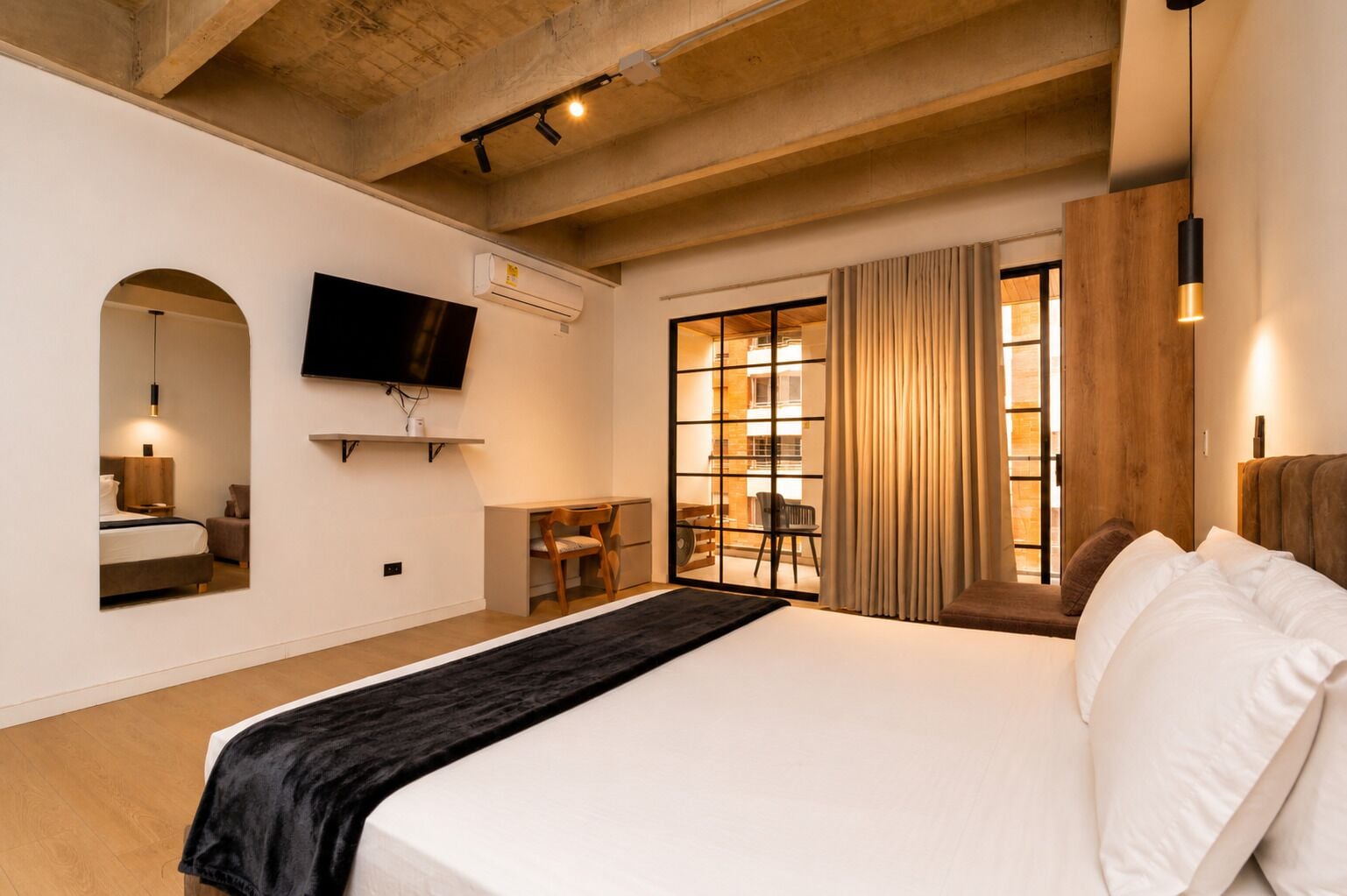 A hotel room with a large bed, a flat-screen TV mounted on the wall, a desk with a chair, and a window with curtains.
