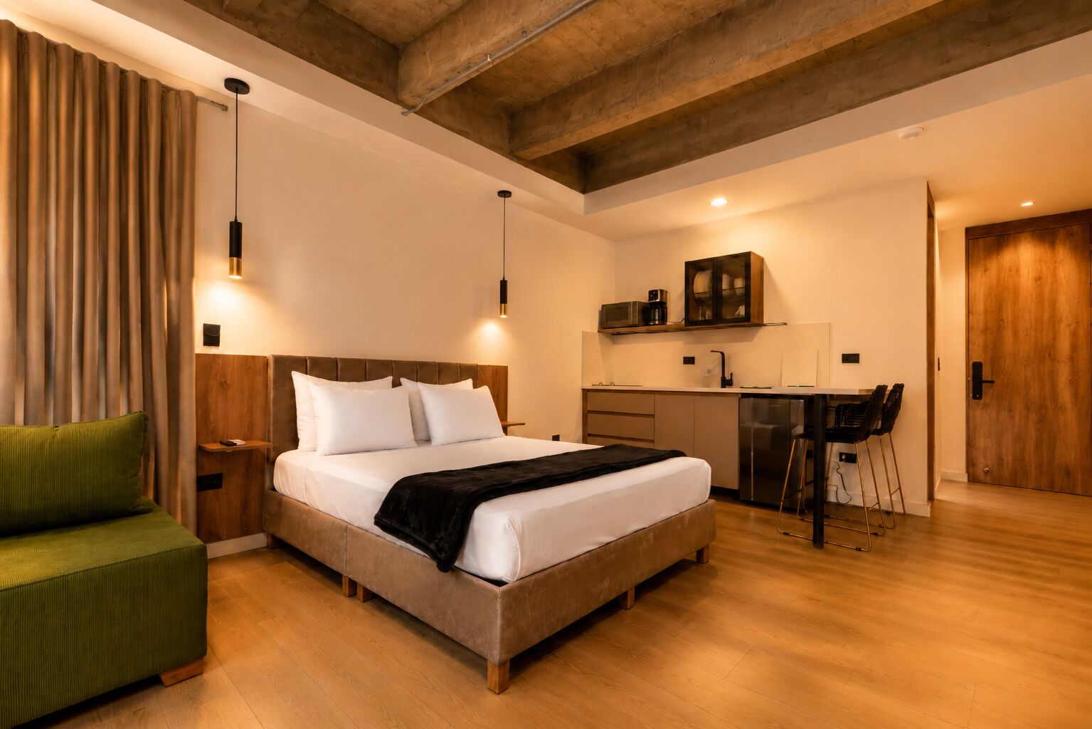 Ein modernes Schlafzimmer mit einem großen Bett, Nachttischen, einem grünen Sessel, einer Holzbalkendecke und einer Küchenzeile.