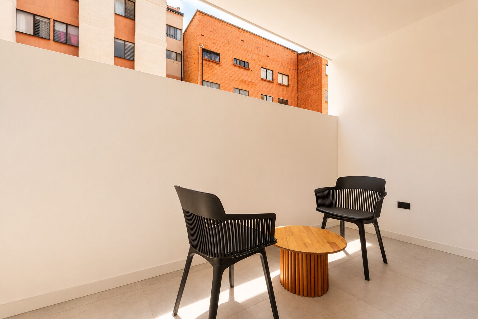 A modern interior with two black chairs and a wooden table, a white wall, and a view of a brick building.