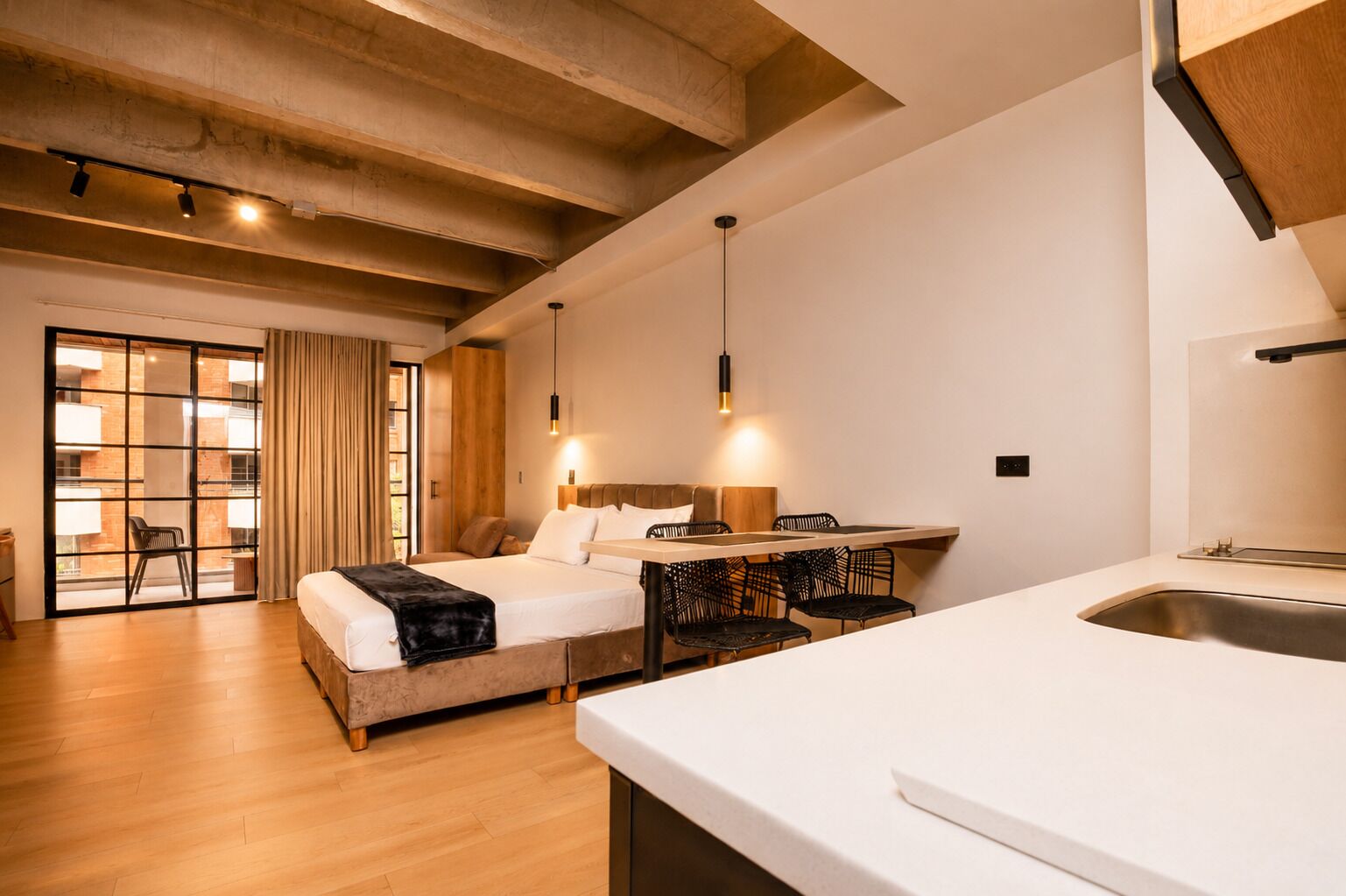 Une chambre d’hôtel moderne dotée d’un grand lit, d’un bureau avec des chaises, d’un coin cuisine et d’une porte-fenêtre coulissante donnant sur un espace extérieur.