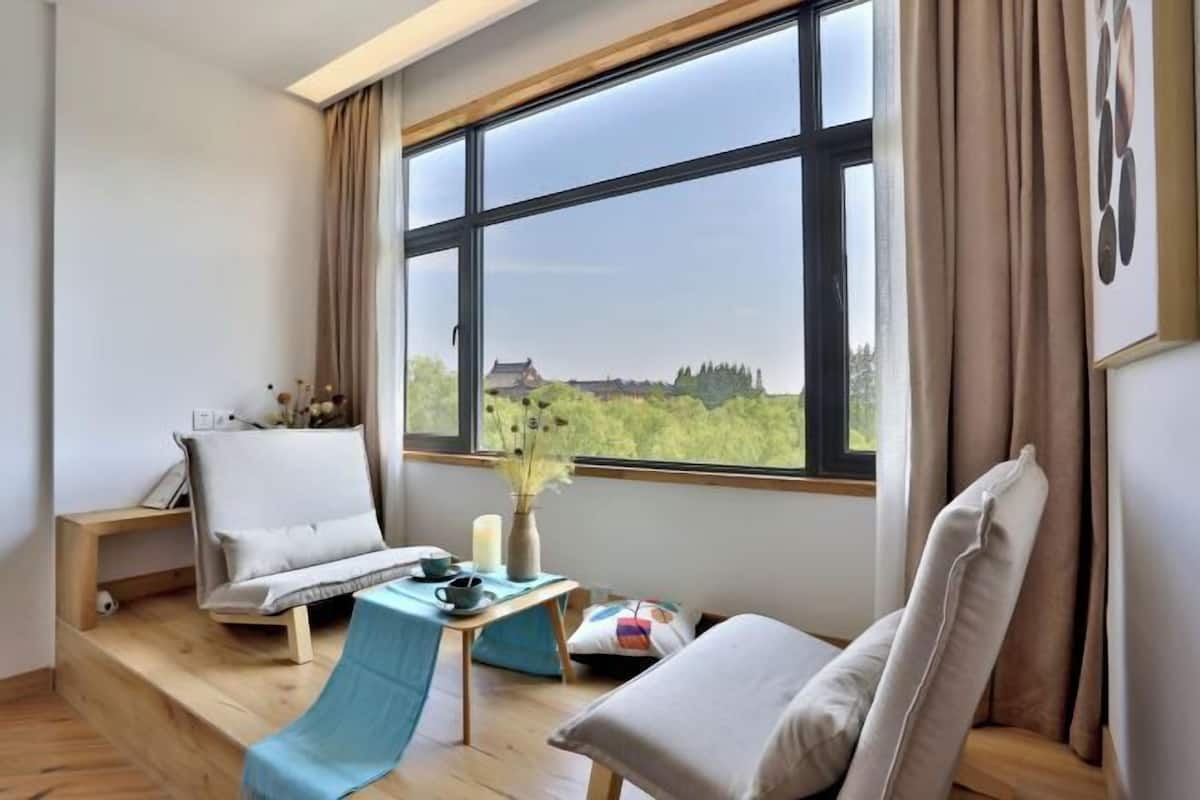 Ein Zimmer mit großem Fenster, einer Couch, einem Sessel, einem kleinen Tisch und einer Vase mit Blumen.
