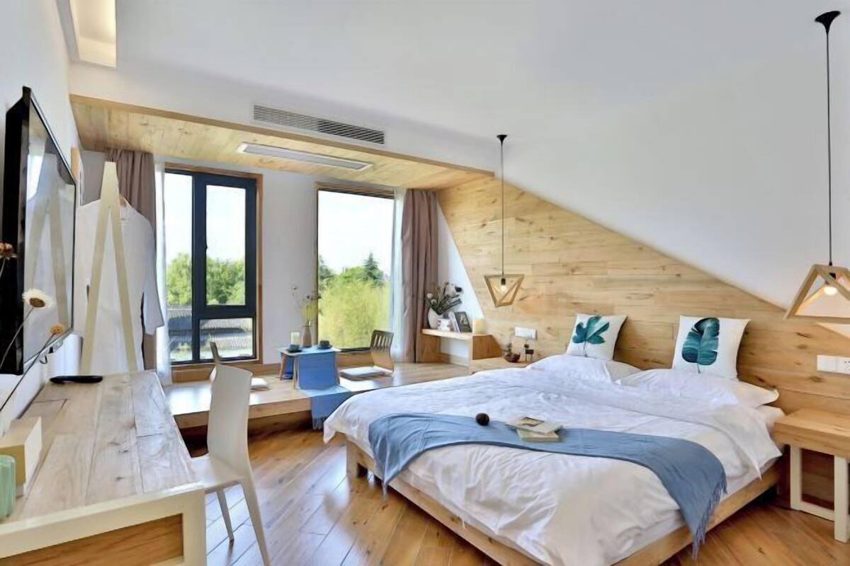 Ein modernes Schlafzimmer mit schrägen Wänden, einem großen Bett, einem Schreibtisch und einem Fenster mit Blick ins Grüne.